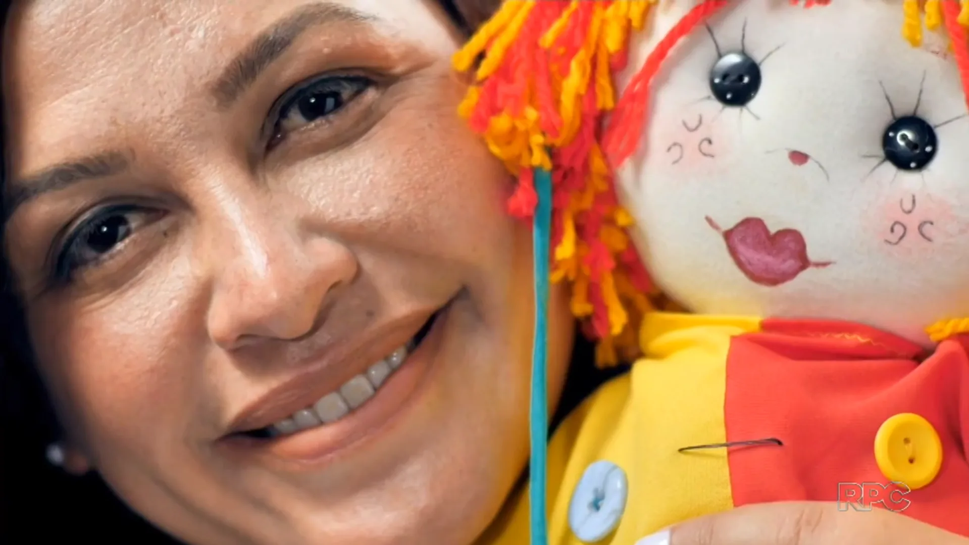 Tia Edi sorrindo ao lado de uma boneca artesanal, em reportagem da RPC.